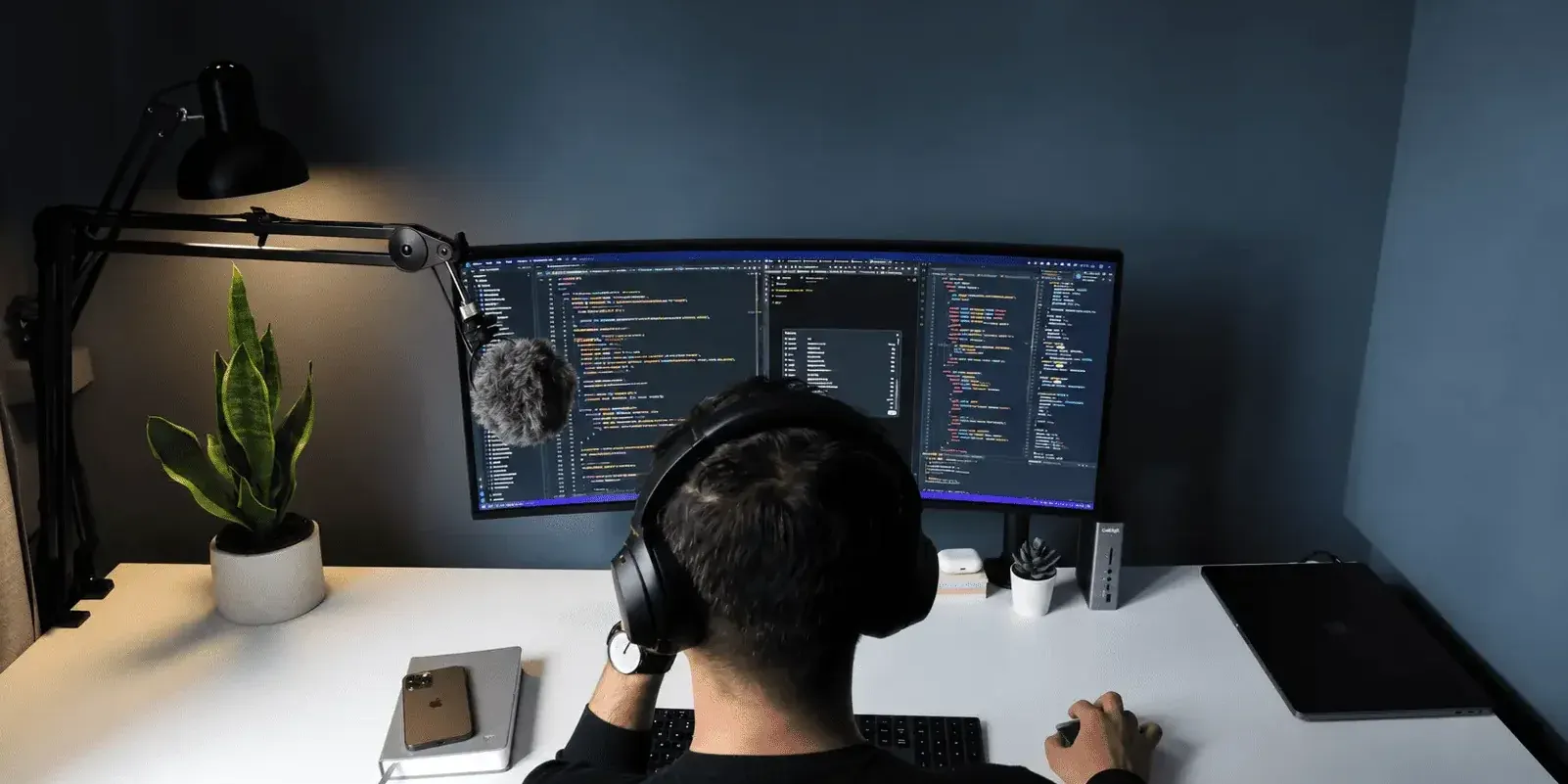 Developer working late at a dark desk setup, viewing code on a curved dual-monitor display, wearing headphones with a studio microphone nearby